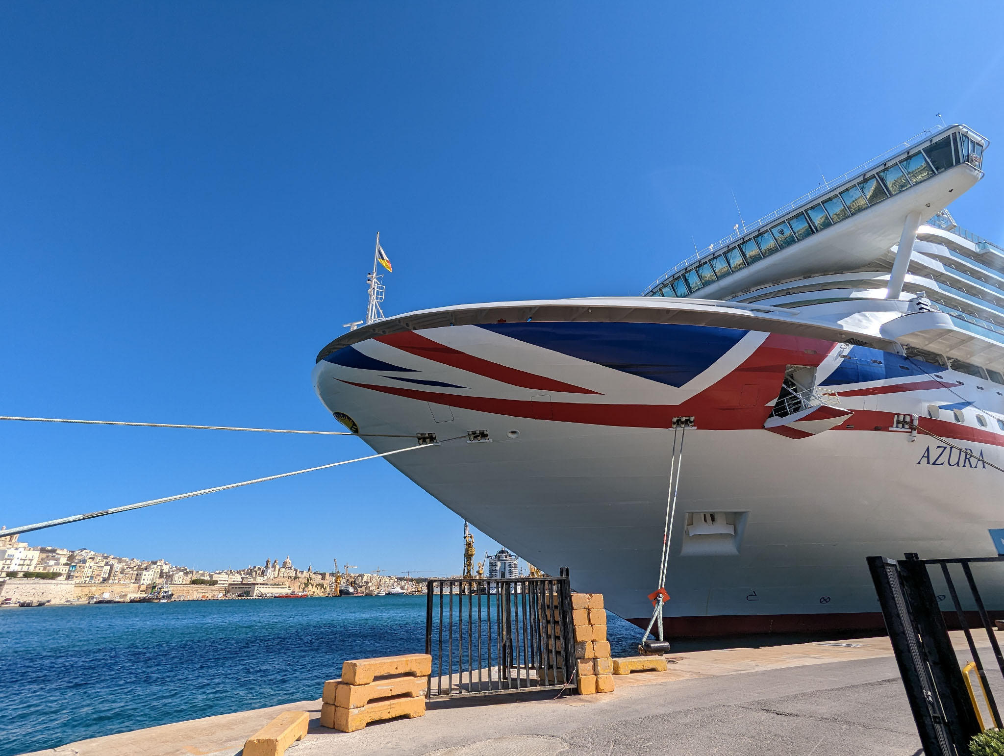 A cruise ship docked at port; we're close to it with its prow decorated with a Union Flag and the word Azura on it. A bright blue sky and sunshine above.