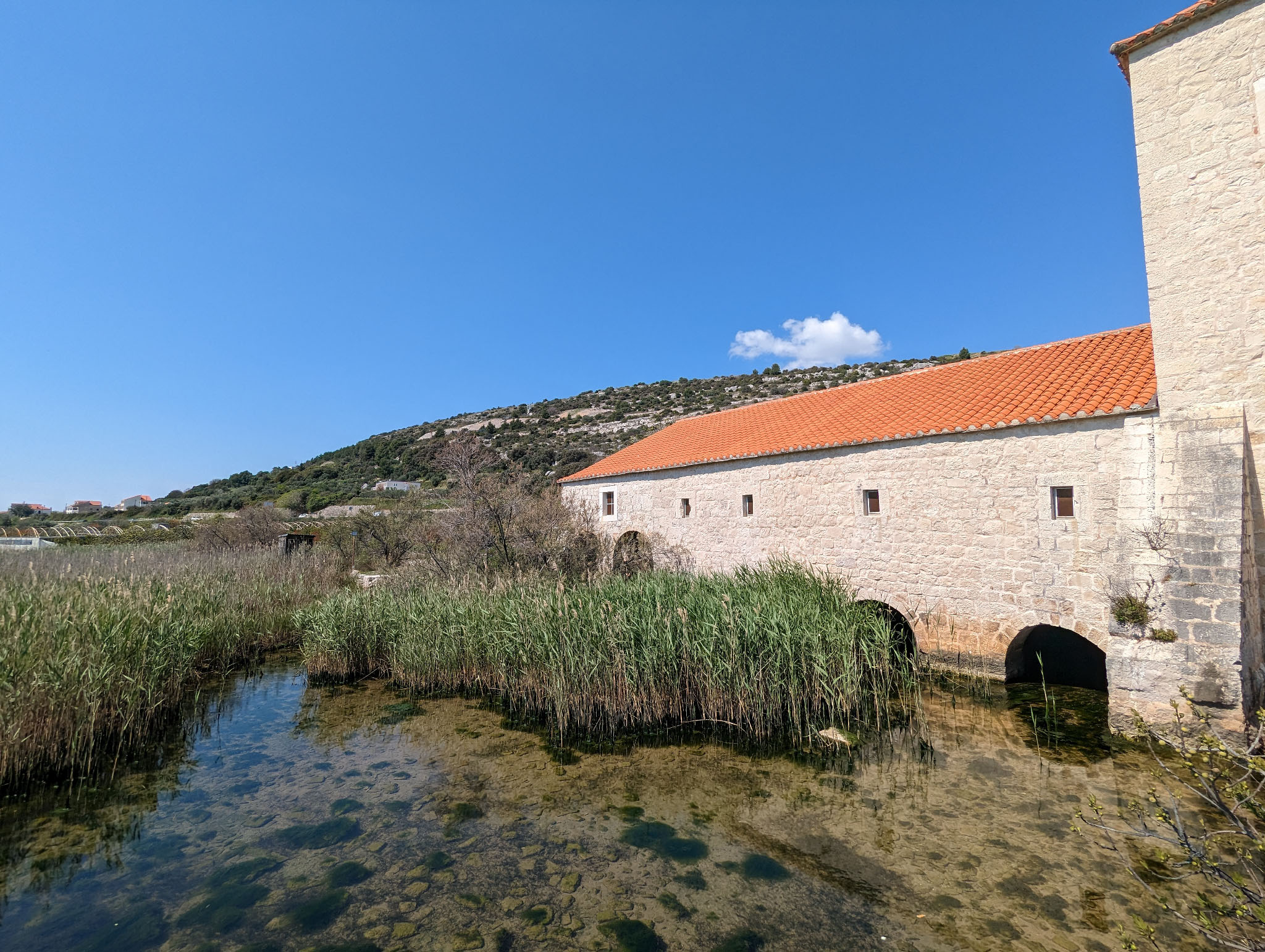 Rear of a mill with stone walls, some arches in the base where the lake water flows, and some red tile roofing. Reeds grow in the water. A single small cloud is in a bright blue sky.