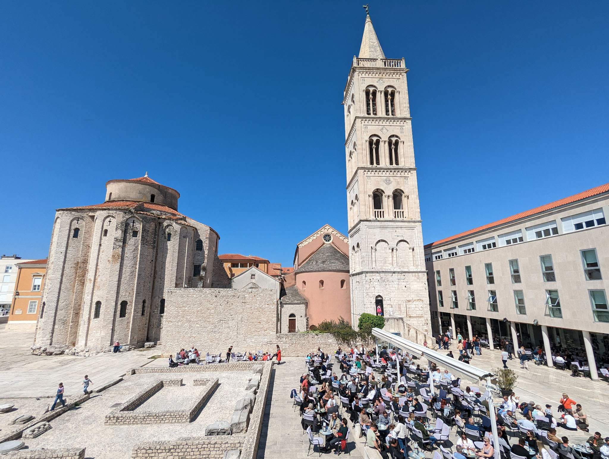 A medieval square showing signs of Roman ruins in the foreground to the left, and masses of people sat at tables right of that, enjoying drinks and food in the sunshine. Behind, a medieval church building and tall tower frame the sunny shot.