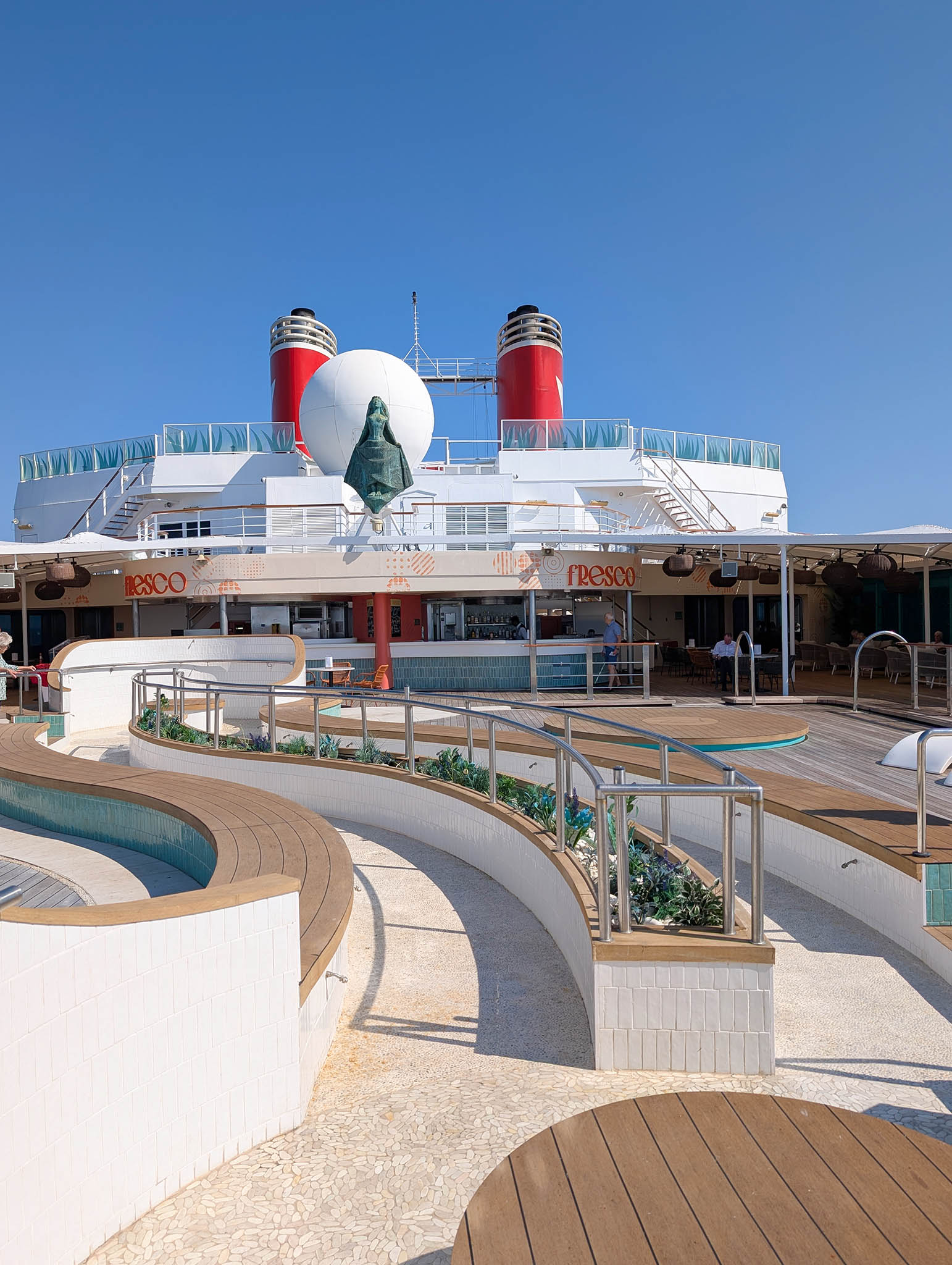 Aft deck space on a cruise ship on a sunny day with a curving, walking structure filling the space as we look forward to a bar and pizza area and two red funnels.