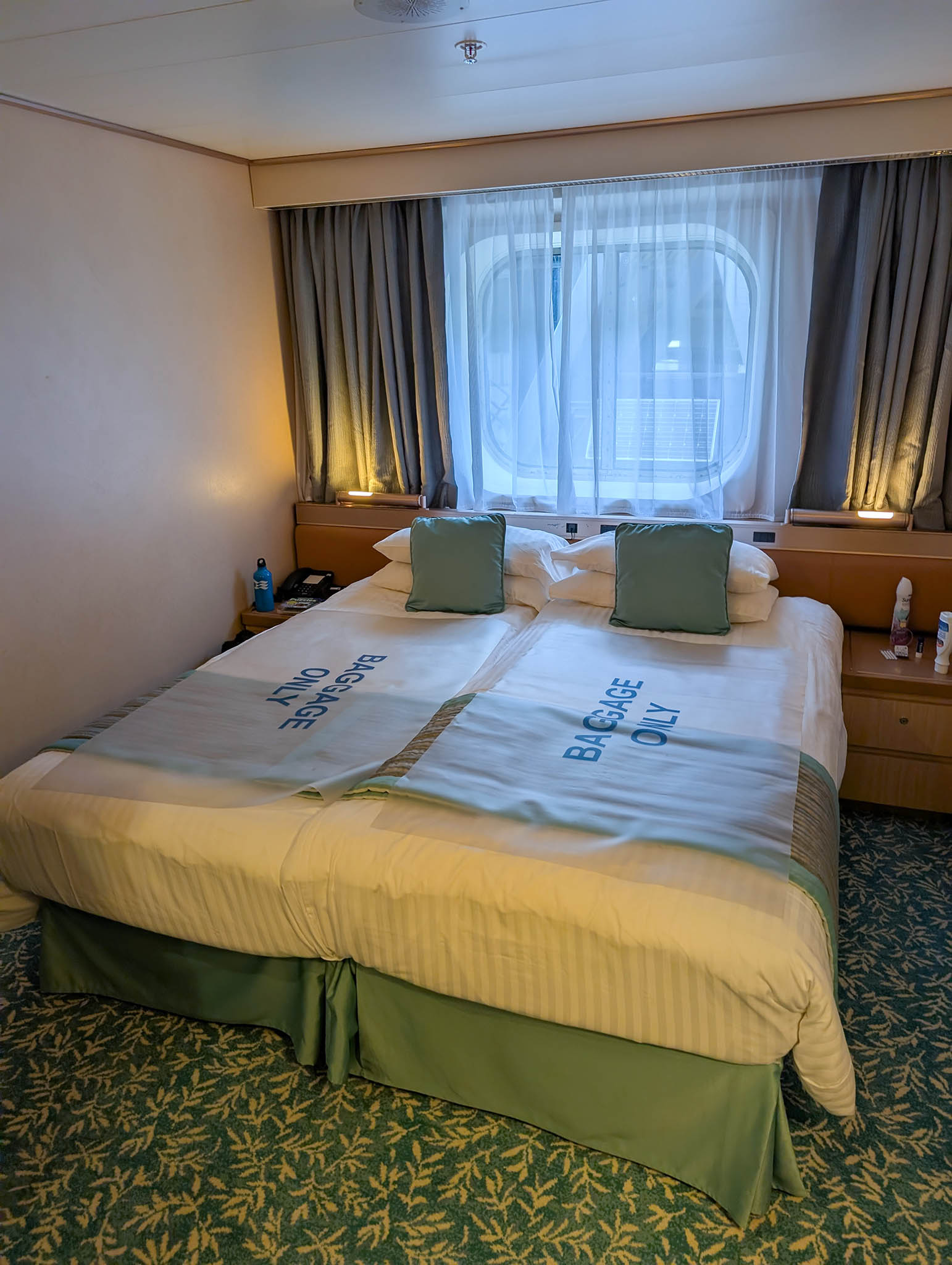 A cruise ship cabin with rounded square window set in the external wall above and behind a bed arranged in Queen configuration.