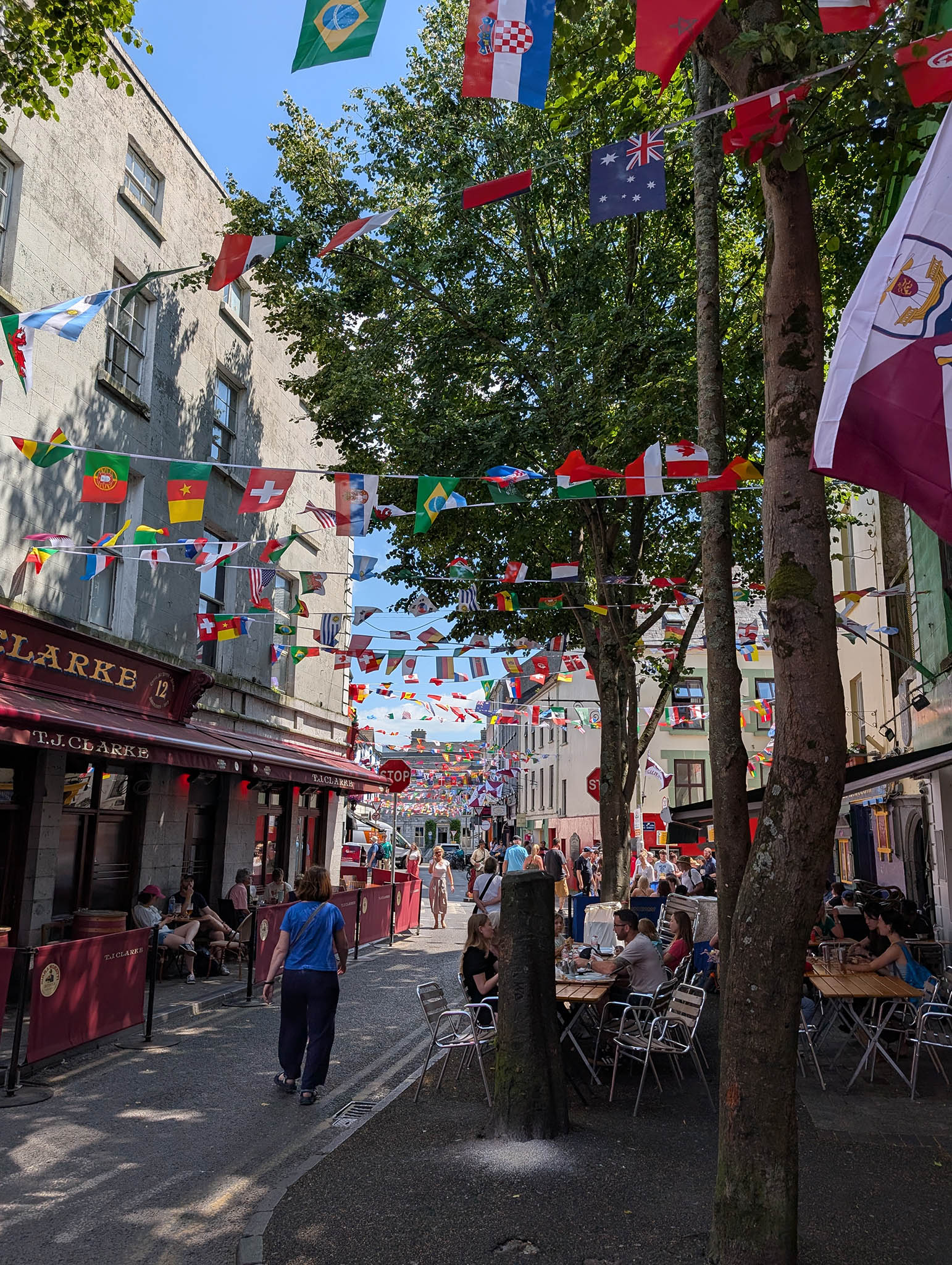 A sunny day and a busy, narrow, winding street of bars and cafes, shaded by close buildings and some trees, with national flags on bunting all down the street as the trip coincides with the World Cup.