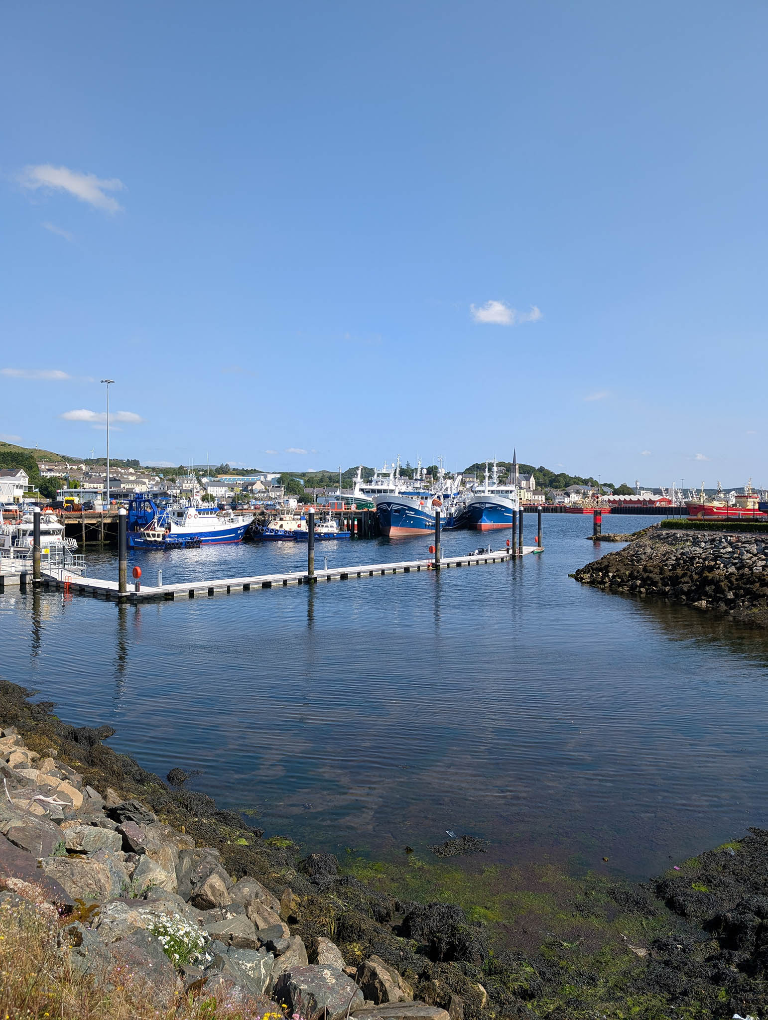 A fishing port with several trawlers docked a pontoons and piers. Crystal clear water and an almost cloudless sky overhead.
